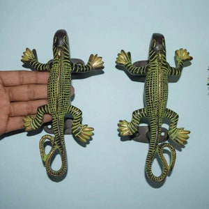 Pode incluir: Um par de puxadores de porta decorativos em forma de lagartos verdes e pretos com caudas enroladas. Os puxadores são de metal e têm um acabamento desgastado.