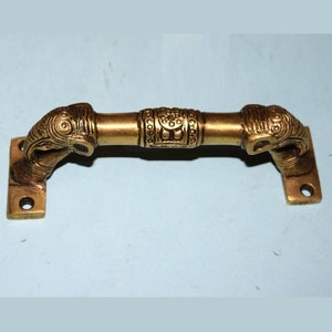 May include: A brass door handle with an ornate design featuring two elephant heads on either end. The handle is mounted on two rectangular plates.