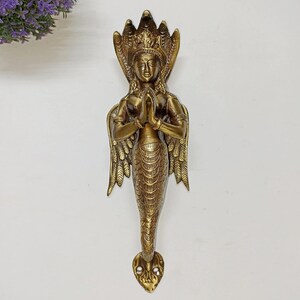 Può includere: Una maniglia per porta in metallo color ottone raffigurante una figura con busto umano, ali e coda di serpente. La figura ha le mani giunte in posizione di preghiera e un cappuccio di cobra a più teste dietro la testa. La base ha due fori.