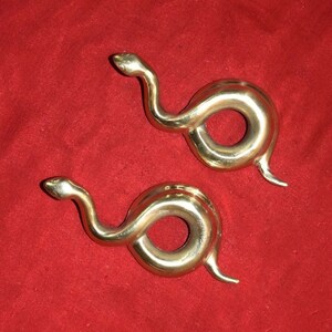 May include: Two brass snake-shaped napkin rings. Each ring features a coiled body with a raised head and a curved tail. The rings are displayed on a red background.