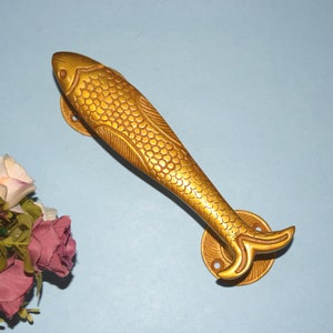 Może przedstawiać: Złoty, rybi uchwyt do drzwi z detalicznym wzorem. Uchwyt jest wykonany z metalu i ma gładkie, polerowane wykończenie.
