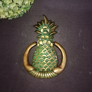 Könnte beinhalten: Ein goldener Ananas-Türklopf mit einem grün bemalten Ananas-Design. Der Türklopfer hat einen Ring zum Klopfen.