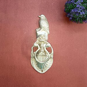 May include: A silver door knocker with a bird design. The knocker is ornate and has a detailed design. The bird is perched on a base with a ring for knocking.