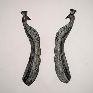 May include: Two decorative peacock-shaped handles. The handles are made of a dark metal with intricate detailing. The peacock's body is curved, with a textured pattern and gold accents. The head features a crown and a beak.