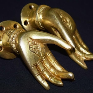 May include: A pair of gold-colored metal door handles shaped like human hands. Each handle features a circular mounting plate with four screw holes, a wrist band detail, and an engraved floral symbol on the palm of the hand.
