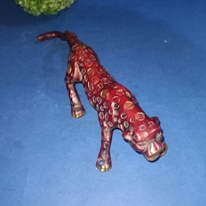Peut inclure: Une petite figurine de léopard en métal rouge et or. Le léopard est en position accroupie, la tête tournée sur le côté. La figurine est détaillée avec des taches et des rayures.
