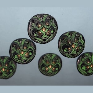 May include: Six round, black and green buttons with a raised, detailed design of a face with gold accents.