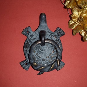 May include: A dark bronze-toned metal door knocker shaped like a turtle with an intricate embossed shell pattern. A circular, twisted metal ring is attached to the centre of the shell, serving as the handle for this functional hardware piece.