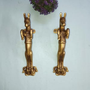 May include: A pair of antique brass door handles, each in the shape of a stylized donkey. The handles are a warm gold colour, with detailed carvings on the donkey's body and base. They are mounted on a light blue wall, with a purple plant above.