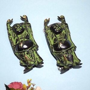 Op de afbeelding: Twee groene en zwarte metalen deurkloppers in de vorm van lachende Boeddha-figuren met opgeheven armen. De figuren dragen gewaden en hebben gouden accenten.