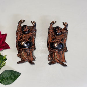 以下が含まれることがあります： 腕を上げた笑顔の仏陀の茶色の金属製置物2つ。置物は白い表面の上に立っています。