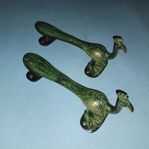 Może przedstawiać: Dwa uchwyty do drzwi w kształcie pawia z zielonymi i złotymi akcentami. Uchwyty są wykonane z mosiądzu i mają tradycyjny design.