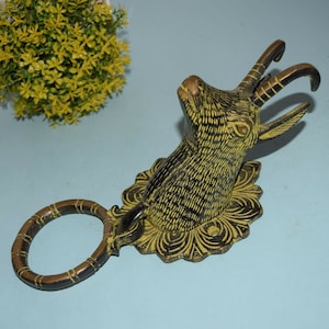 Könnte beinhalten: Ein Türklopfer aus Messing in Form eines Ziegenkopfes mit einem Ringgriff. Der Ziegenkopf ist mit schwarzer und gelber Farbe verziert und hat ein florales Design um die Basis.