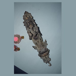 Może przedstawiać: Metalowa dekoracja ścienna w kolorze ciemnobrązowym z rzeźbą przedstawiającą bóstwo, bogato zdobioną i z motywem kwiatowym. Rzeźba jest zamontowana na ścianie z jasnoniebieskim tłem.