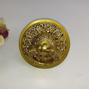 Puede incluir: Un llamador de puerta de metal dorado con un diseño intrincado que presenta un marco circular con un patrón floral detallado y un anillo central con una figura de dragón.