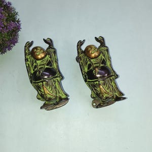以下が含まれることがあります： 腕を上げた緑と金の笑い仏の置物2つ。置物は金属製で、使い込まれたような仕上げになっている