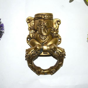 May include: A gold-toned brass door knocker featuring a seated Ganesha figure with a decorative ring. The knocker is intricately detailed with floral and geometric patterns.