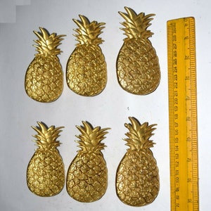 May include: Six gold-colored pineapple-shaped decorative items. The pineapples are all the same size and shape, and they are arranged in two rows of three. The pineapples are on a white background.