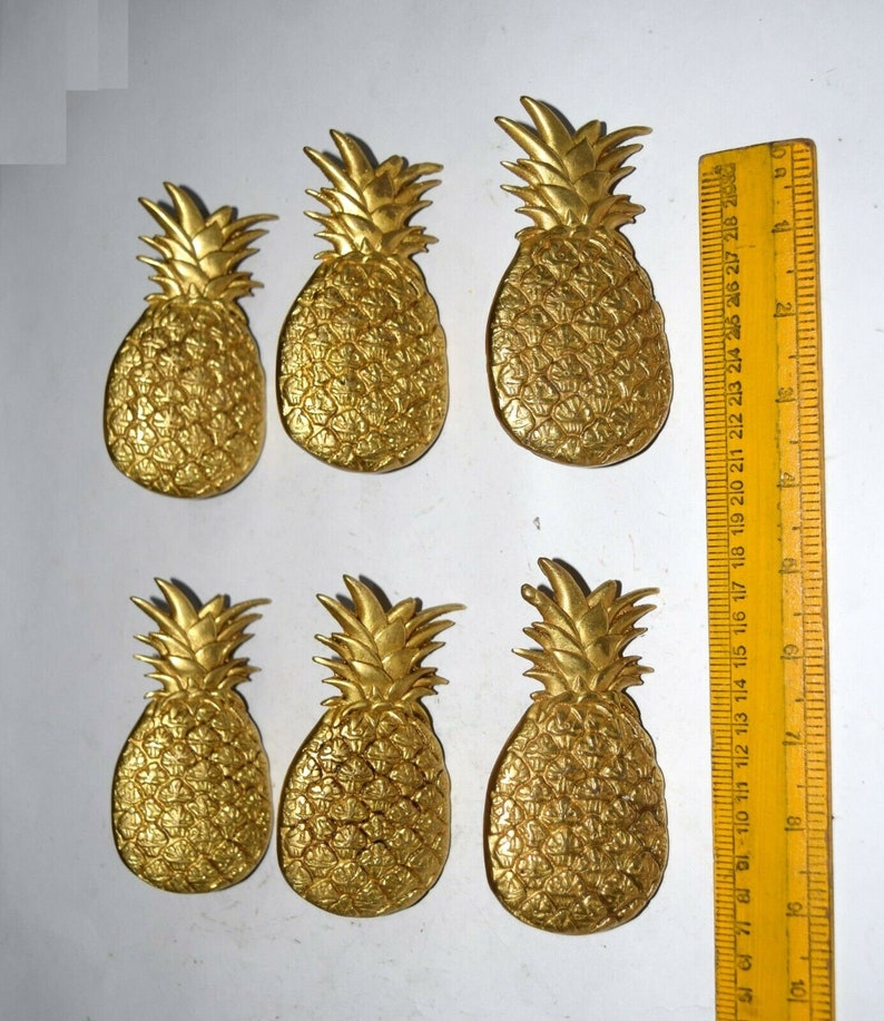 May include: Six gold-colored pineapple-shaped decorative items. The pineapples are all the same size and shape, and they are arranged in two rows of three. The pineapples are on a white background.