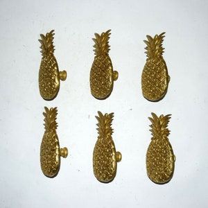May include: Six gold pineapple-shaped cabinet knobs. The knobs are detailed with a textured pineapple design.