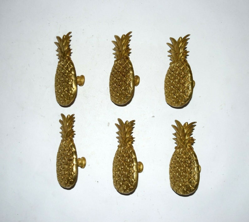 May include: Six gold pineapple-shaped cabinet knobs. The knobs are detailed with a textured pineapple design.
