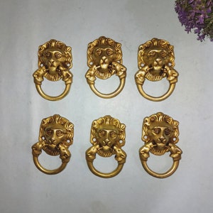 May include: Six gold lion head drawer pulls with a ring handle. Each pull has a detailed lion face with a mane and a ring handle.
