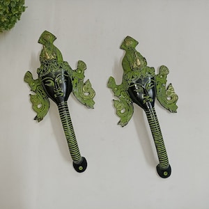 Puede incluir: Dos tiradores de puerta decorativos con acabado verde y negro. Cada tirador presenta una cara estilizada con detalles intrincados. Los tiradores están unidos a una base de metal negro.