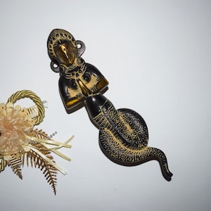 May include: A brass door handle shaped like a woman with a snake tail. The woman is in a prayer position with her hands clasped together. The snake tail is coiled around her legs.