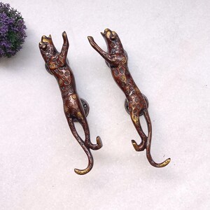 May include: Two bronze-colored leopard-shaped door handles. The handles are detailed with spots and are in a climbing pose, with their front paws raised. The handles are mounted on round, dark-colored plates.