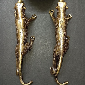 Könnte beinhalten: Zwei bronzefarbene dekorative Wandhaken in Leopardenform. Die Haken sind mit Flecken versehen und befinden sich in einer Kletterpose. Die Haken sind aus Metall und auf einer dunkelgrauen Oberfläche montiert. Oben befindet sich eine grüne Pflanze.