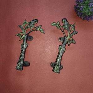 Puede incluir: Un par de tiradores de puerta de metal negro y verde con forma de árboles con loros posados en las ramas.