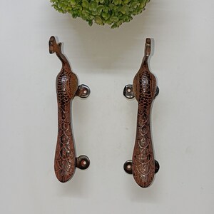 Może przedstawiać: Dwa brązowe metalowe klamki do drzwi w kształcie pawia. Klamki mają szczegółowy wzór i są zamontowane na białym tle.