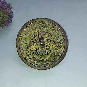 Puede incluir: Una tapa redonda de metal de estilo antiguo con un diseño detallado de dragón. La tapa es principalmente dorada con acentos verdes y marrones, con intrincados patrones calados y un asa central.