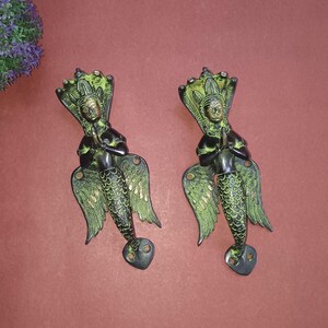 May include: A pair of decorative door handles made of brass. Each handle features a winged figure with a human head and a fish tail. The figures are in a praying position with their hands clasped together. The handles are painted in shades of green and black.