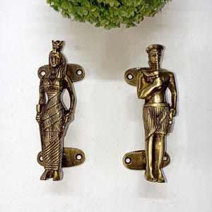 Peut inclure: Deux poignées de porte antiques en laiton, chacune représentant un pharaon égyptien. Les poignées sont montées sur des plaques rectangulaires avec des trous de vis. Les figures sont en position debout.