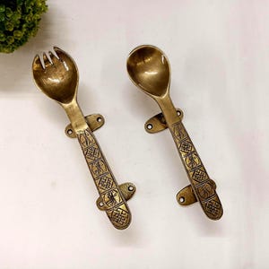 Puede incluir: Dos utensilios de servir de latón antiguo: un tenedor y una cuchara. El tenedor tiene tres púas, y ambos utensilios tienen mangos decorativos con un patrón grabado. Cada pieza tiene una placa de montaje con dos agujeros.