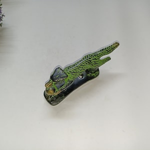 Puede incluir: Un llamador de puerta de metal verde y negro con forma de animal agachado con un acento dorado.