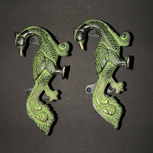 Może przedstawiać: Dwa metalowe haczyki ścienne w kształcie pawia, w kolorze zielonym i czarnym, z złotymi akcentami. Haczyki są zaprojektowane tak, aby wyglądały jak pawie z rozłożonymi ogonami.