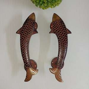 Könnte beinhalten: Ein Paar dekorative Türklinken in Fischform. Die Griffe sind aus Messing und haben eine dunkelbraune Oberfläche mit goldenen Akzenten. Die Fische haben Schuppen und Flossen.
