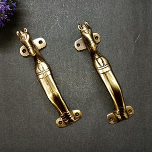 May include: Two antique brass door handles with a horse head design. The handles are a warm gold colour and have a detailed, vintage aesthetic. Each handle is approximately 18 cm long.