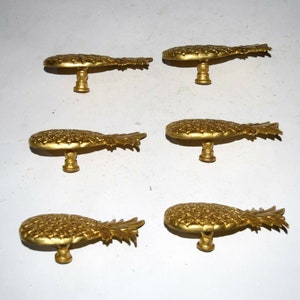 May include: Six gold pineapple-shaped cabinet knobs with a detailed design. The knobs are all facing the same direction and are arranged in two rows of three.