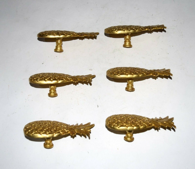 May include: Six gold pineapple-shaped cabinet knobs with a detailed design. The knobs are all facing the same direction and are arranged in two rows of three.