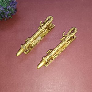 May include: Two gold-toned door handles shaped like alligators. The handles are detailed with textured scales and have a simple, elegant design.