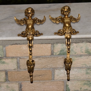 Könnte beinhalten: Zwei goldene Engel-Strumpfhalter mit filigranen Details. Die Engel halten ihre Flügel ausgestreckt und haben ein cherubinisches Gesicht. Die Halter sind an einem dekorativen Rollendesign befestigt.