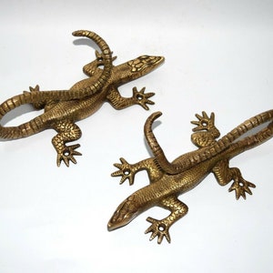 May include: Two gold-toned metal lizard wall hooks. Each lizard has a curled tail that forms a hook.