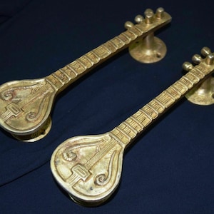 Puede incluir: Un par de tiradores de puerta de color dorado con forma de sitar, un instrumento musical tradicional de la India. Los tiradores tienen un diseño detallado y están hechos de metal.