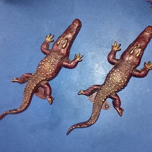 以下が含まれることがあります： 赤と金の仕上げが施された、装飾的なワニのドアノブ2個。ノブは金属製で、リアルなワニのデザインです。