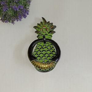 May include: A green and black pineapple door knocker with a gold ring. The pineapple is detailed with scales and a gold crown.