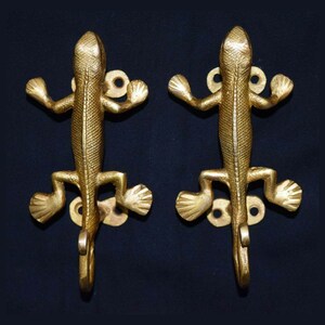 May include: Two gold-toned metal lizard-shaped hooks. The hooks are mounted on a dark background.