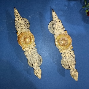 May include: Two ornate gold door handles with intricate floral designs and a central circular element. The handles are made of metal and have a polished finish.